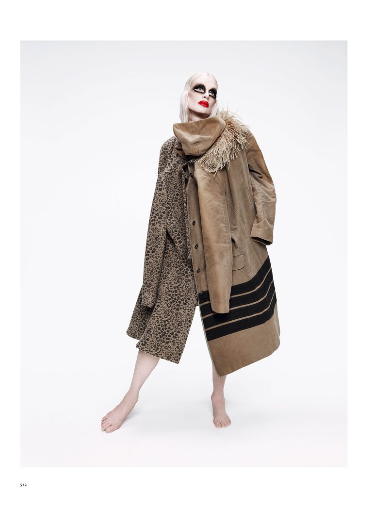Fashion shot of an adult model in oversized leopard print and beige coats with heavy dark makeup and bare feet against a white backdrop.