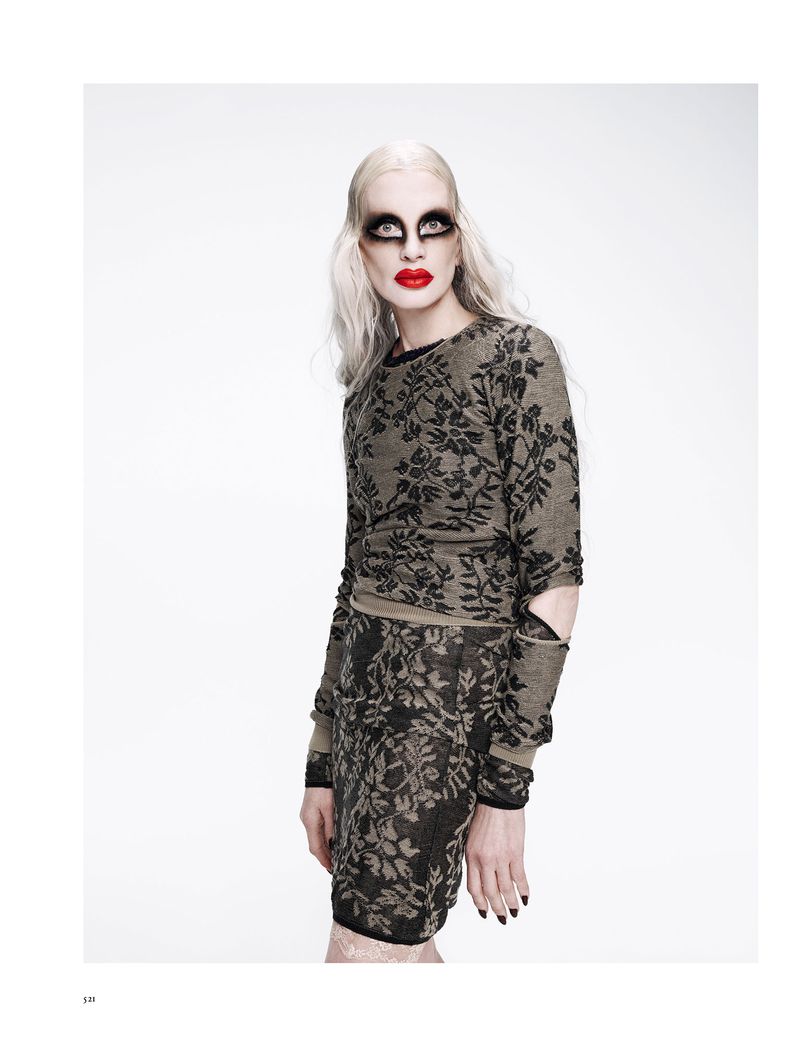 Fashion shot of a model with dramatic makeup, wearing a patterned sweater and skirt. Against a bright white background in studio lighting.