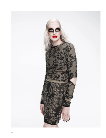 Fashion shot of a model with dramatic makeup, wearing a patterned sweater and skirt.  Against a bright white background in studio lighting.