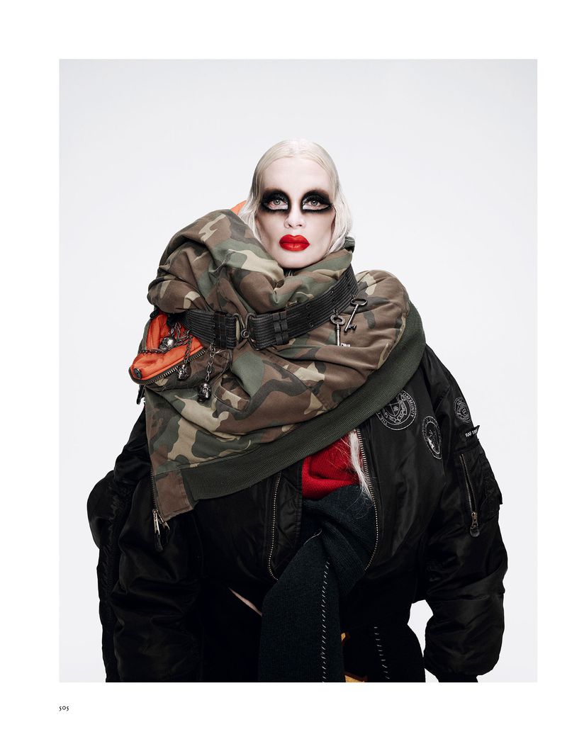 A female fashion model with dramatic eye makeup and red lipstick wears a camouflage jacket and black bomber jacket against a white backdrop.