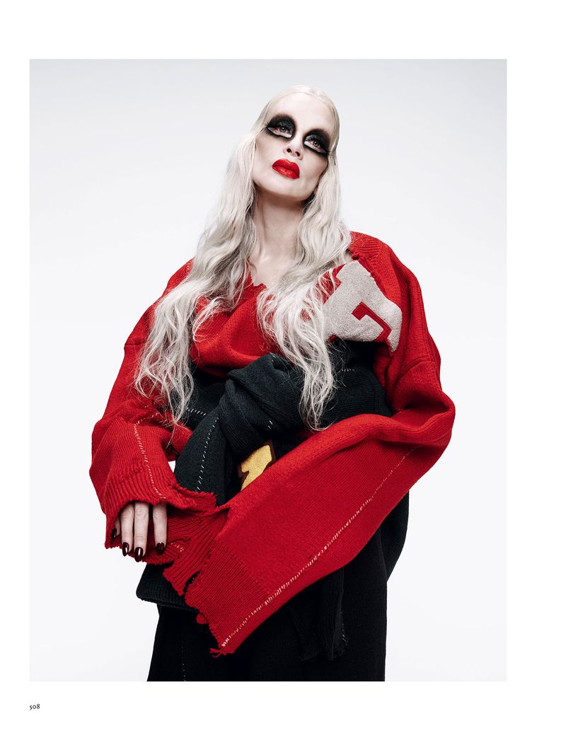 A pale woman with long white hair wearing a red sweater and heavy black makeup looks upward against a white background in a fashion style portrait.