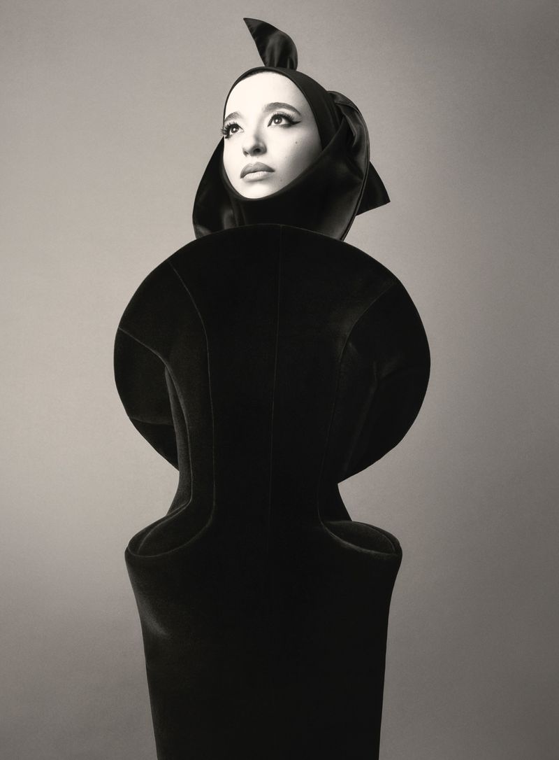 High fashion portrait of a woman wearing a black sculptural hood and dress in front of a muted gray backdrop with dramatic studio lighting.