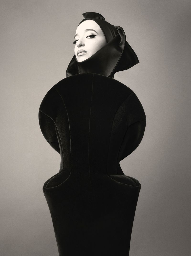 Monochrome studio portrait of an adult female fashion model wearing a dramatic black garment and matching head covering against a grey background.