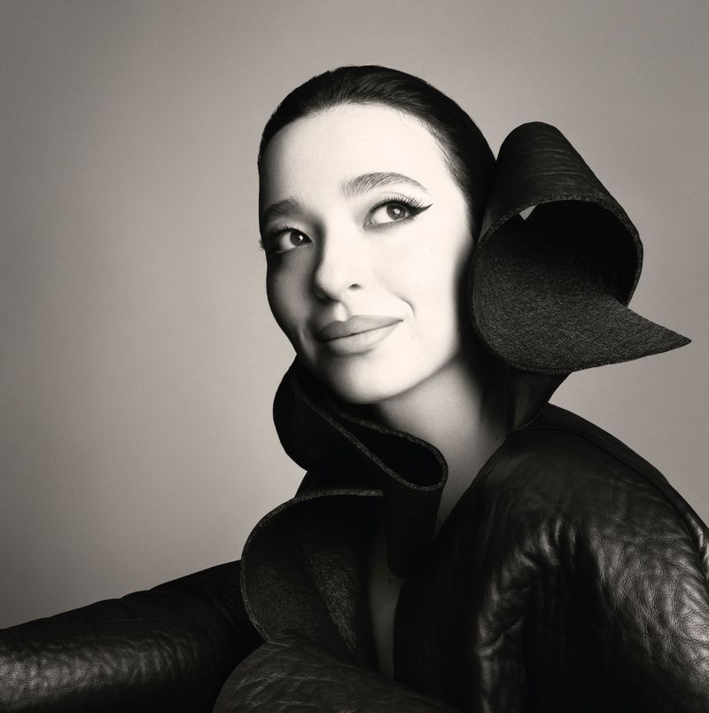 Close-up portrait of a smiling adult woman with dark hair and stylized eye makeup wearing a dramatic black collar and jacket indoors.