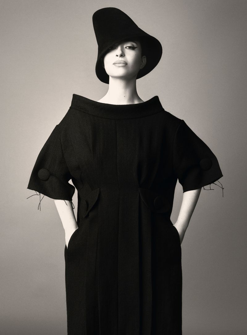 Fashion model wearing an elegant black dress and a matching sculptural hat poses in a studio, looking directly at the camera.