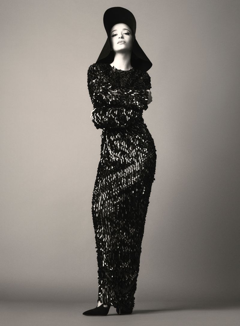 Full-length fashion shot of a woman posing in a long black sequined dress and matching hat against a neutral background.