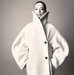 Portrait of Mikey Madison wearing an oversized white coat with a high collar against a neutral studio background