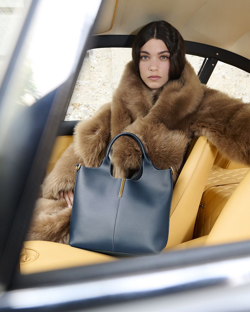 Woman in a fur coat sits in a car holding a blue leather tote bag with gold hardware, creating a fashion editorial moment.