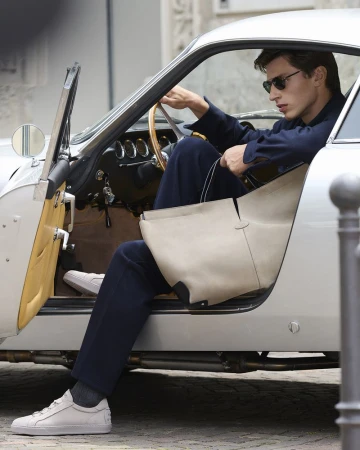 Stylish adult male in sunglasses exiting a classic silver car with a beige leather bag on a city street during daytime.