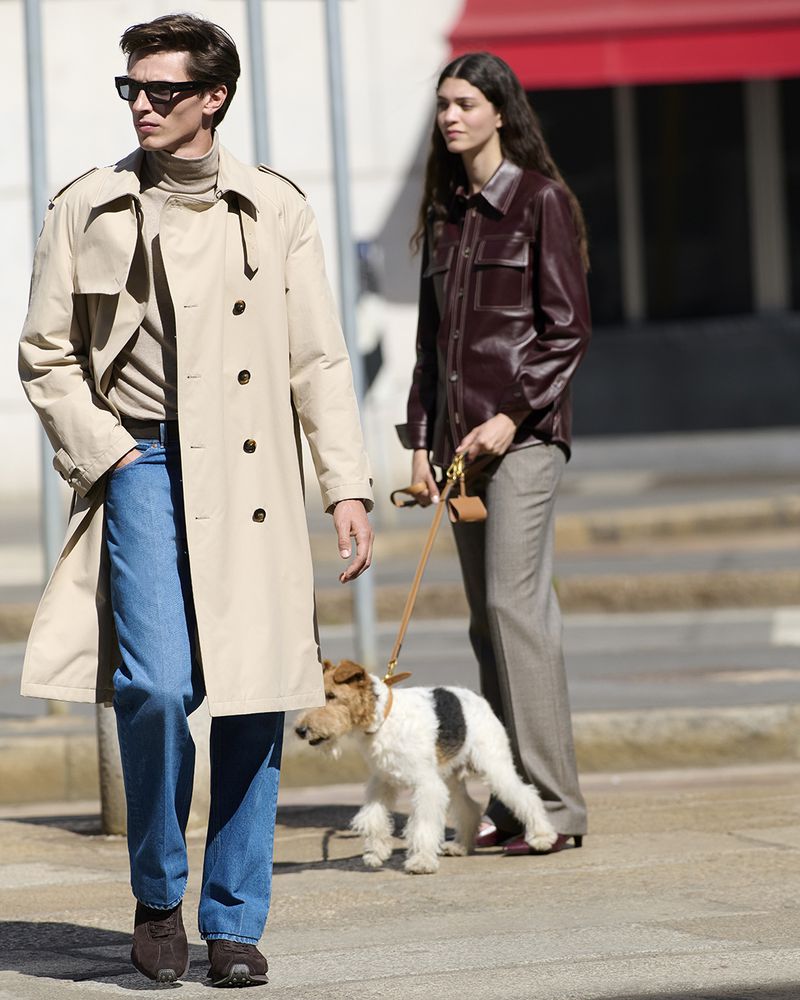 Fashion shot of a stylish man in a trench coat and sunglasses walking with a woman and a dog on a city street on a sunny day.