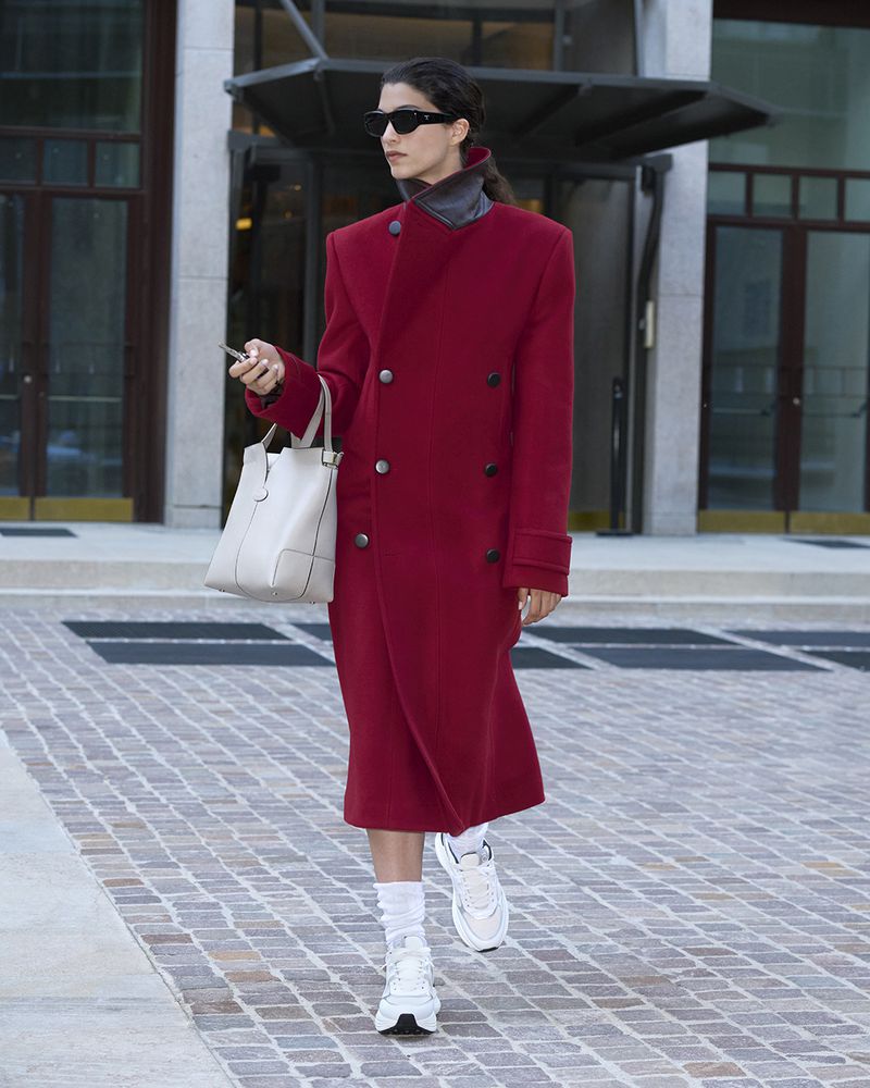 Adult woman in a red overcoat walking outdoors with a white tote bag. She is wearing sunglasses and white sneakers and holding a cell phone.