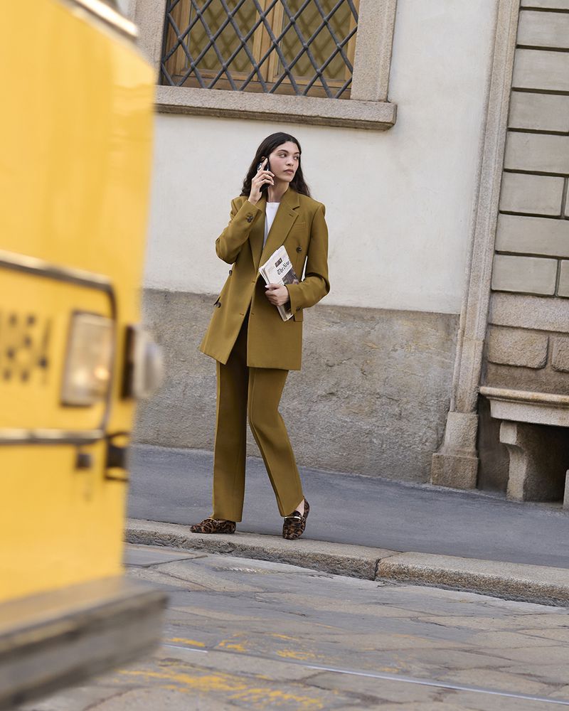 Adult woman in a tailored brown pantsuit is standing on a city street talking on her cell phone near a yellow transit bus.