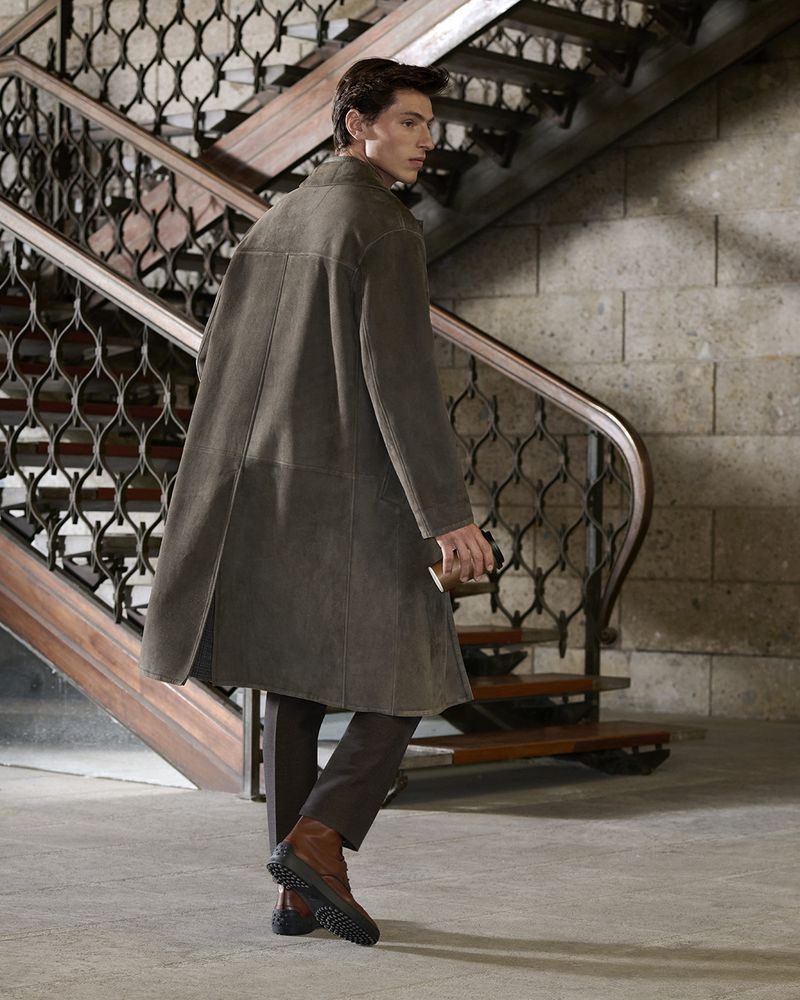 A man walks up a staircase. He's wearing an olive-green overcoat, brown pants, and brown boots. The background shows a winding staircase.