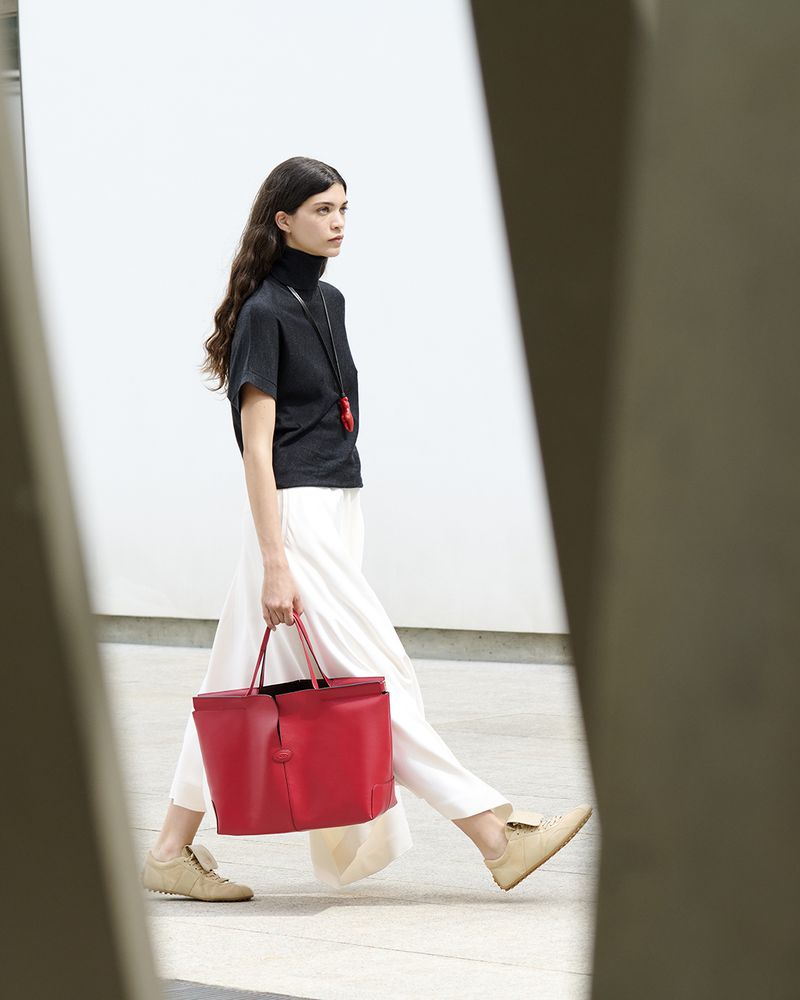 A woman walks with a large red tote bag on a light surface with blurred geometric shapes in the foreground and a neutral background.