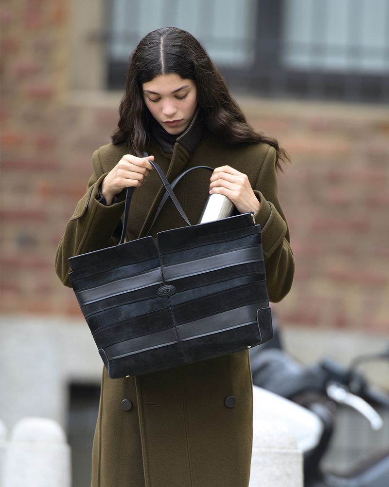 Adult woman with long dark hair, wearing a long olive-green coat, holds a large black handbag outdoors during the day.
