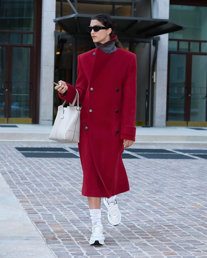 Model wearing a long red double-breasted coat, dark sunglasses, and white sneakers while carrying a light grey tote bag.