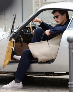A man wearing sunglasses and navy clothing sits in a vintage silver car holding a large beige leather tote bag.