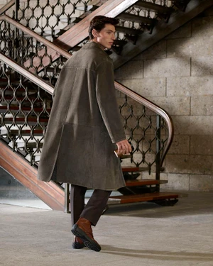 Model wearing a long taupe coat and brown boots descending a grand wooden staircase indoors.