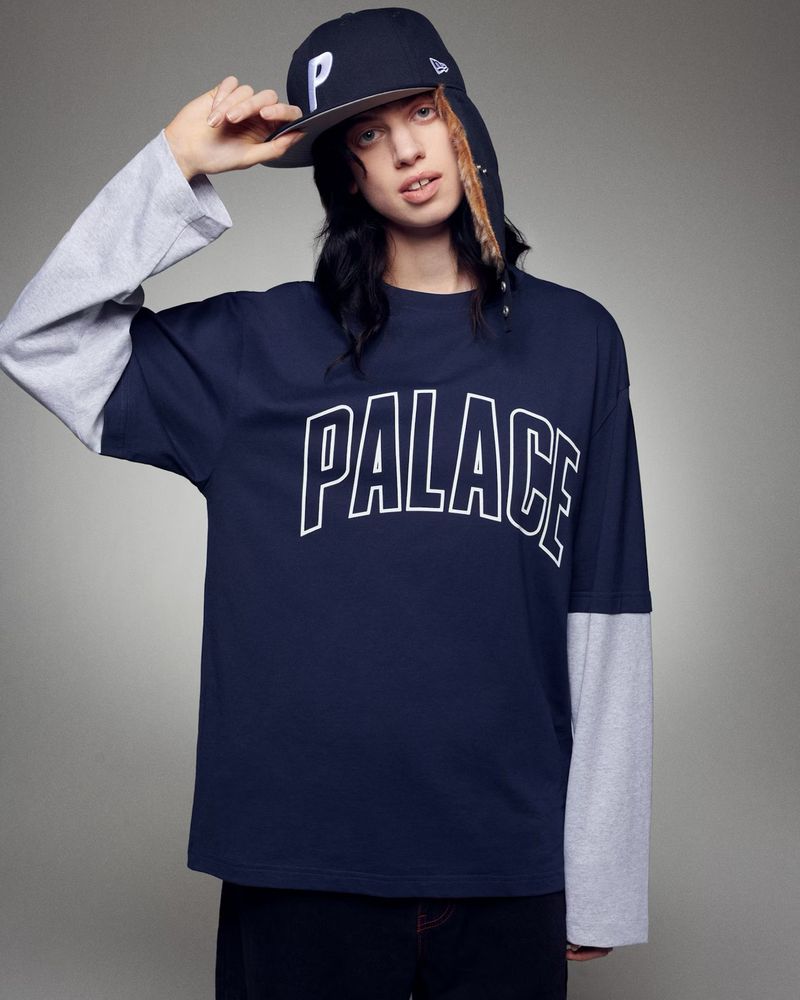 Person wearing a dark blue Palace long-sleeve shirt with a backwards baseball cap, posing indoors against a light gray backdrop.