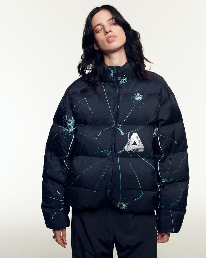Portrait of a woman in a black Palace puffer jacket with broken glass pattern, posing in a studio with a neutral background.