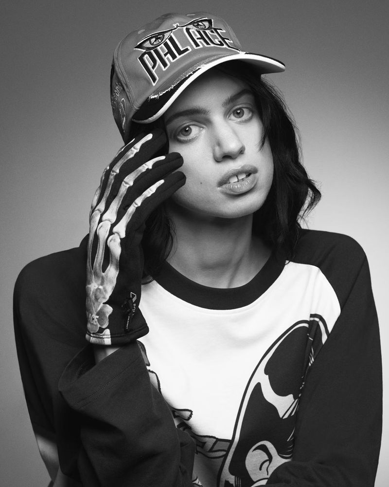 Portrait of a woman wearing a Palace cap and skeleton gloves against a grey background, showcasing a contemporary urban fashion style.