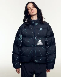 Model Lily McMenamy wearing a black Palace shattered-glass print puffer jacket against a white studio backdrop.