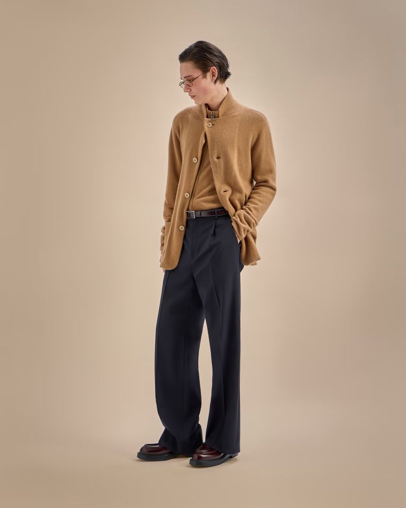 Man in a light brown button-down cardigan, brown shirt, and wide leg dark trousers, standing indoors against a neutral beige backdrop.