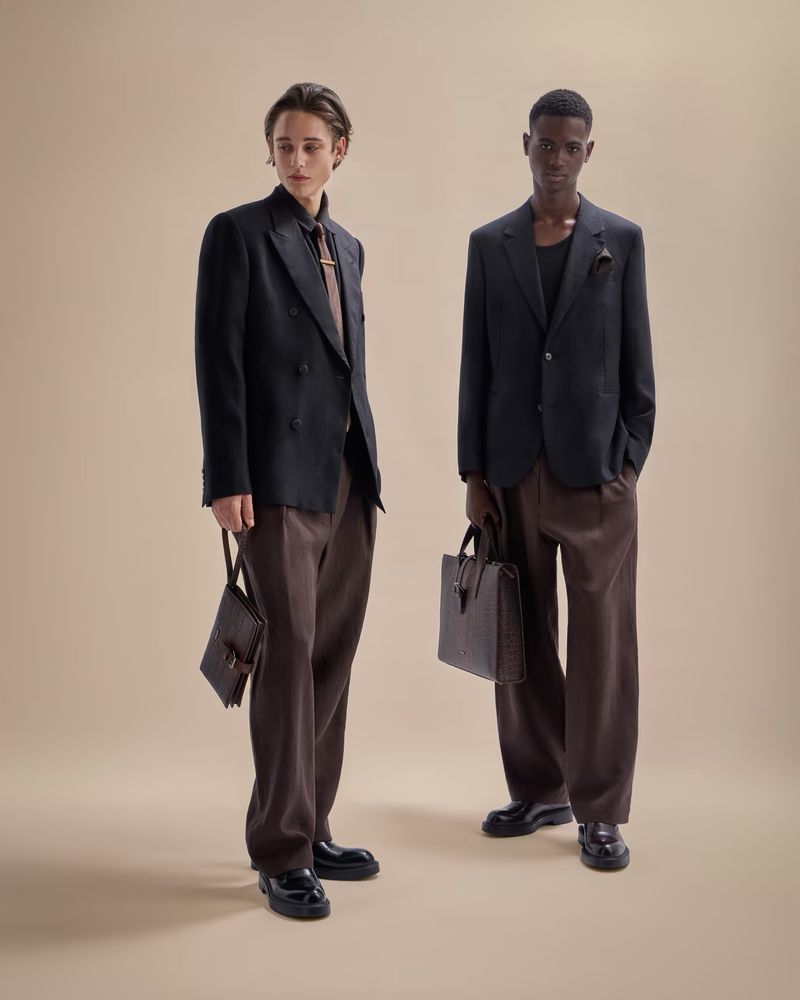 Two male models in dark blazers and brown pants pose indoors on a neutral backdrop holding leather bags, showcasing contemporary fashion.