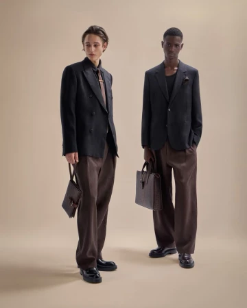 Two male models in dark blazers and brown pants pose indoors on a neutral backdrop holding leather bags, showcasing contemporary fashion.
