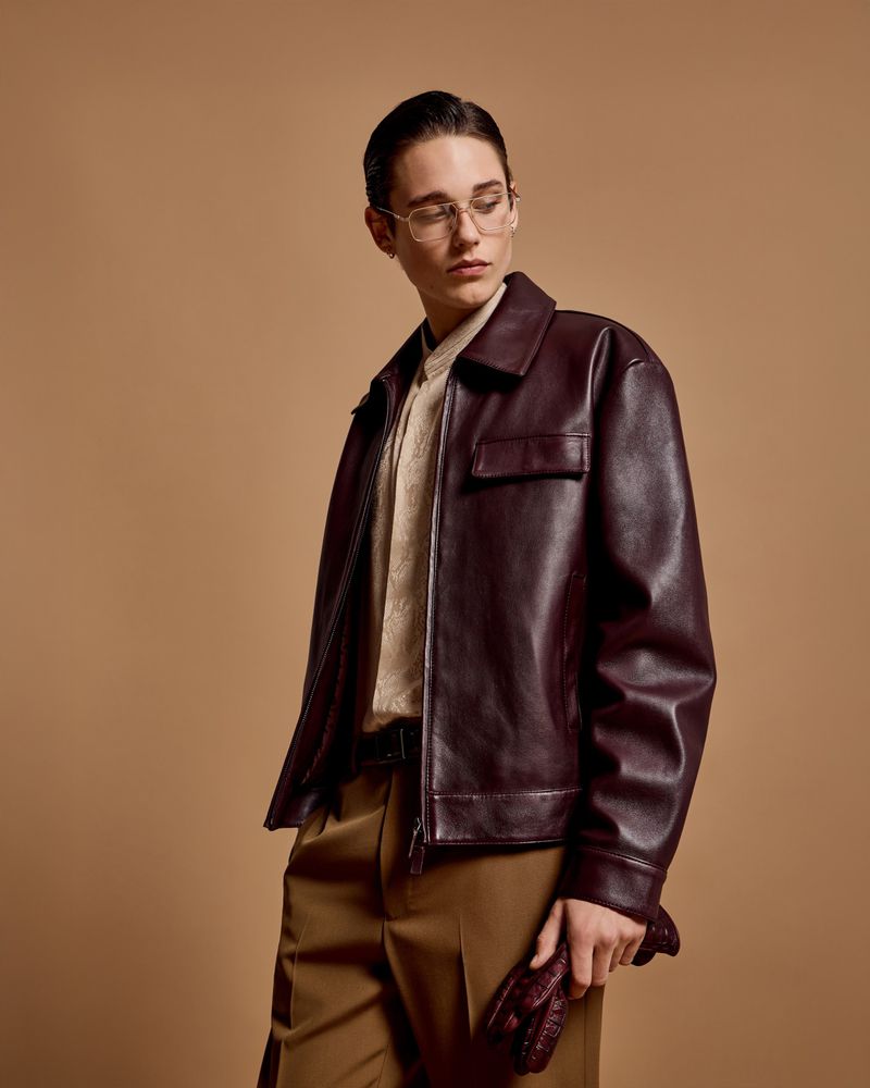 Portrait of a young adult in a brown leather jacket, beige sweater, and brown pants, in a studio with a neutral backdrop.