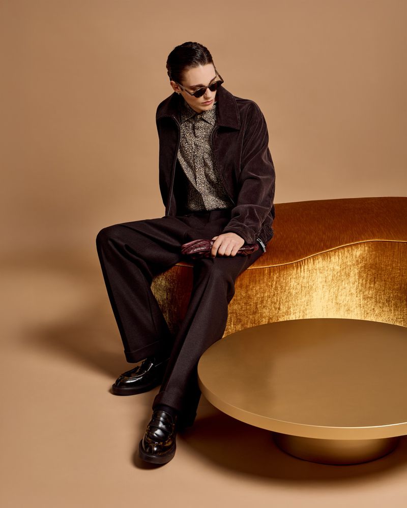 A young adult male model with sunglasses poses on a gold bench in a photo studio wearing corduroy jacket and trousers for fashion editorial.