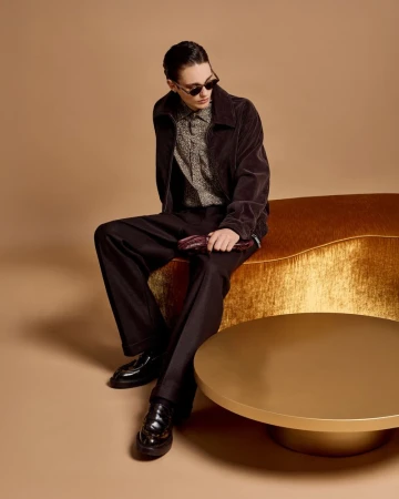 A young adult male model with sunglasses poses on a gold bench in a photo studio wearing corduroy jacket and trousers for fashion editorial.