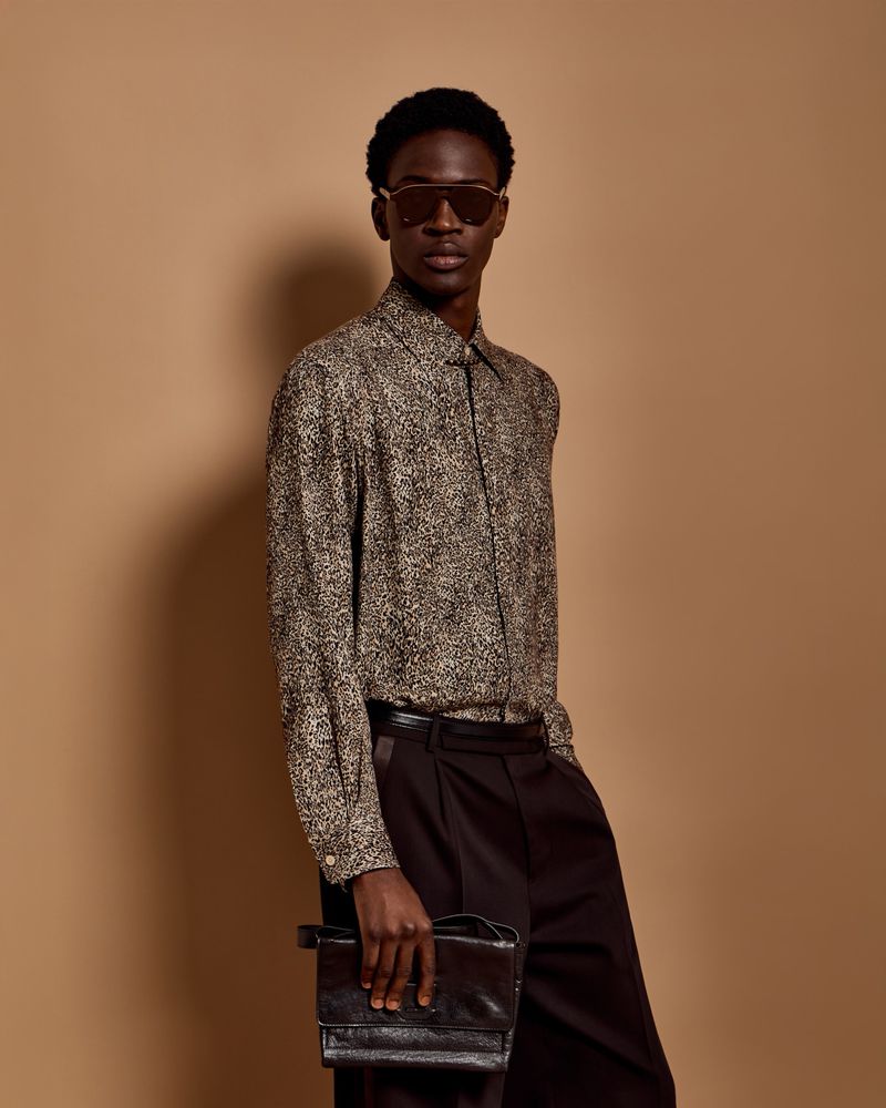 A stylish Black male model poses indoors against a tan backdrop, showcasing a patterned shirt, dark slacks, sunglasses, and a clutch purse.