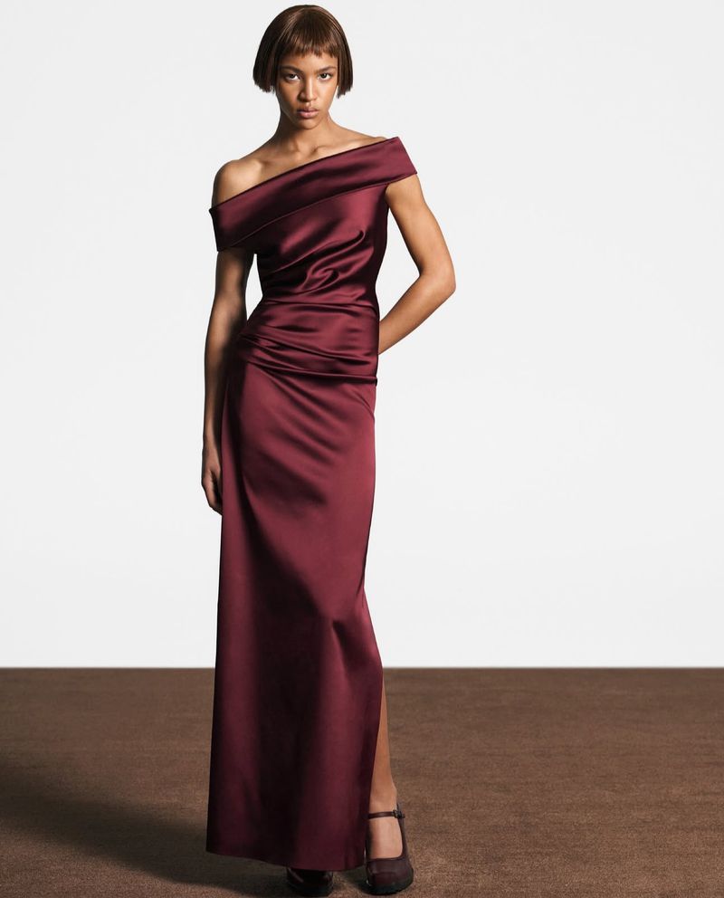A woman in a maroon, off-the-shoulder gown stands in a neutral studio with a brown floor, posing for a fashion editorial shoot.