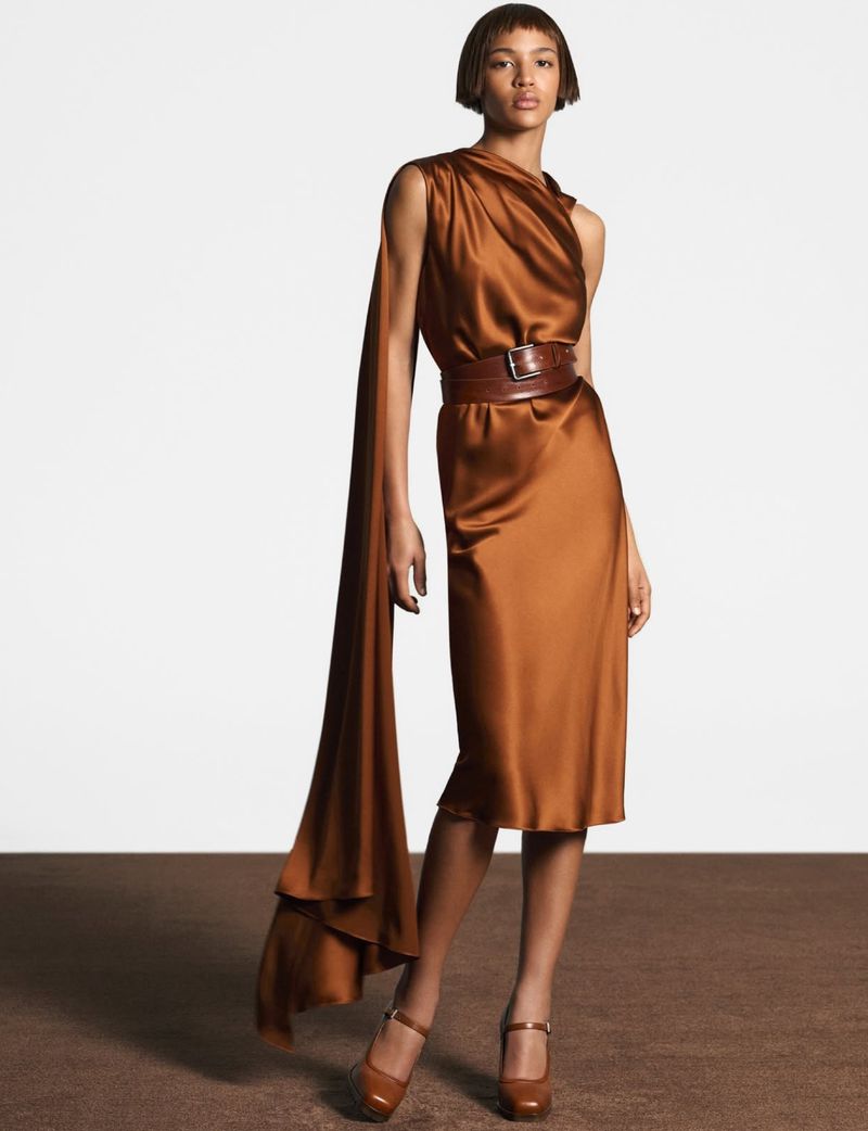 A fashion shot of a woman in a copper-colored dress with brown belt and matching shoes, posing against a white and brown backdrop.