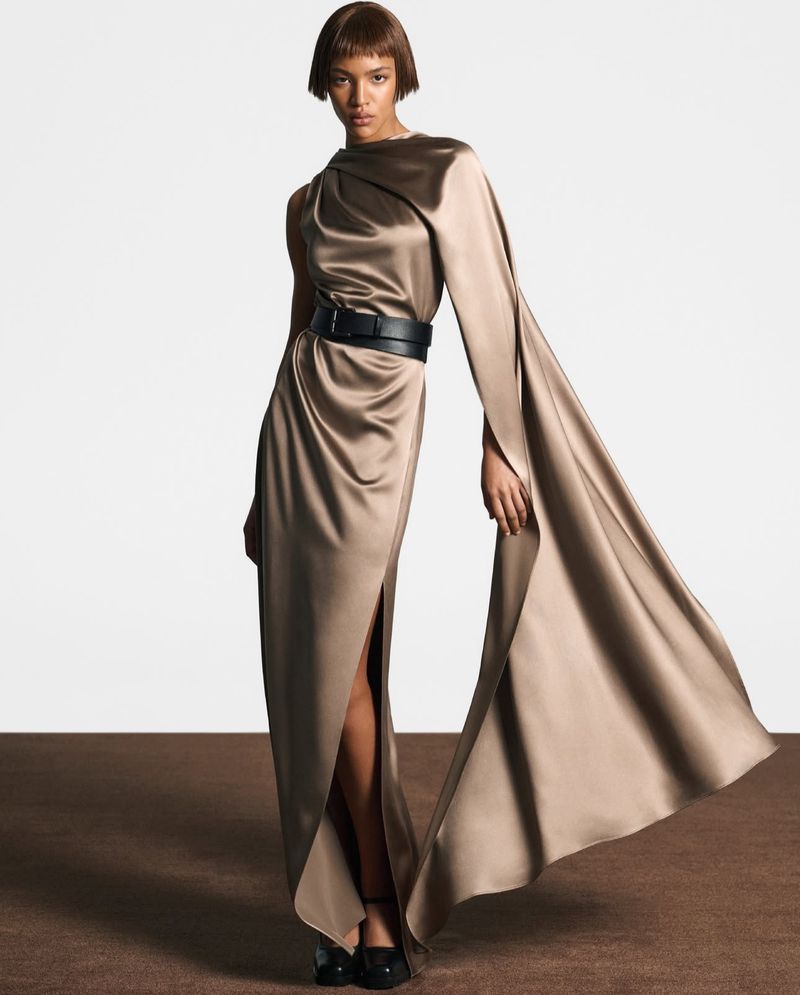Elegant woman in a draped, one-shoulder gown with a black belt and a thigh-high slit stands against a gradient backdrop.