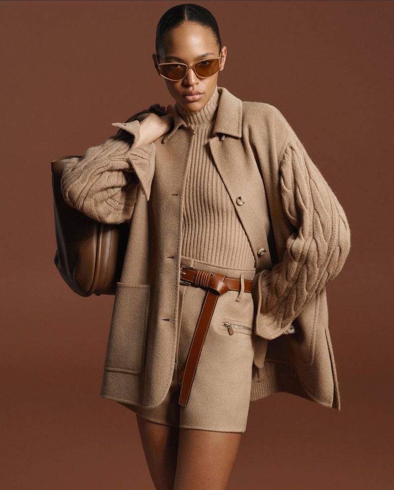 Fashion shot of an adult female model wearing a tan jacket, shorts, and sunglasses, holding a brown purse against a tan background.