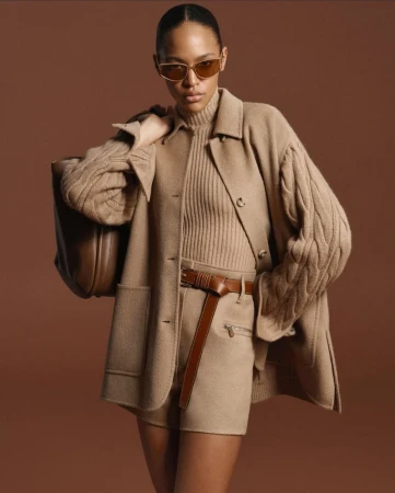 Fashion shot of an adult female model wearing a tan jacket, shorts, and sunglasses, holding a brown purse against a tan background.