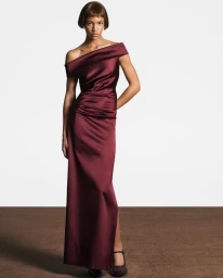 Model Laura Savy in a burgundy satin off-the-shoulder gown standing against a plain backdrop for Max Mara FW25.