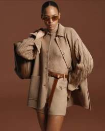 Model wearing a tan coat, matching top, shorts, sunglasses, and holding a brown leather bag against a solid brown background.