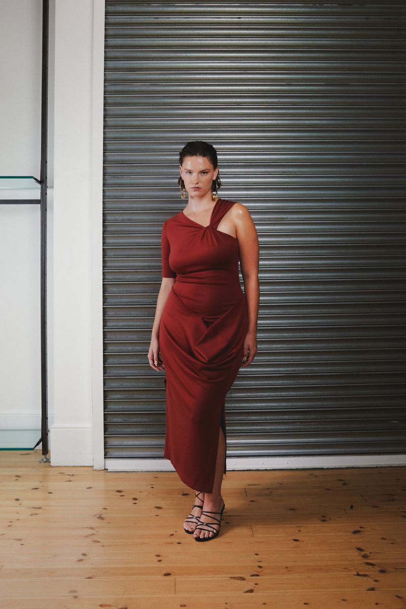 A woman in a burgundy dress poses indoors against a metal roller door. The dress has a unique design, complemented by gold earrings.
