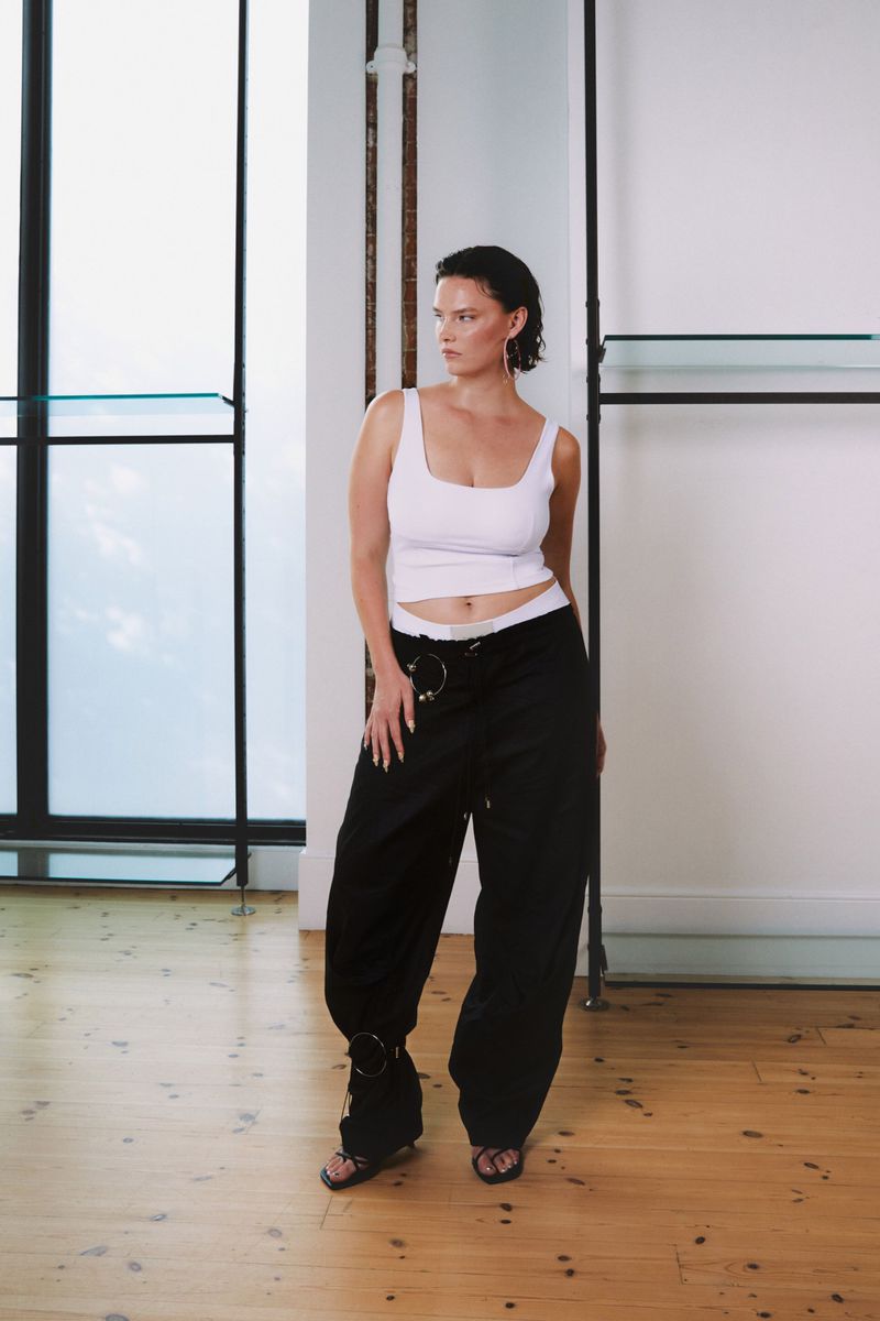 Full-body shot of an adult woman in a white tank top and black pants standing indoors with a neutral expression and posing for the camera.