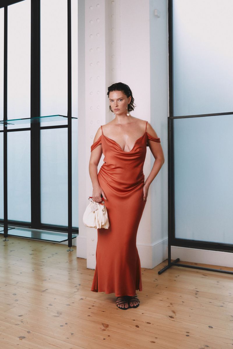 A woman in a rust-colored satin dress and black sandals stands in a minimalist setting with a white handbag, posing for a fashion shot.