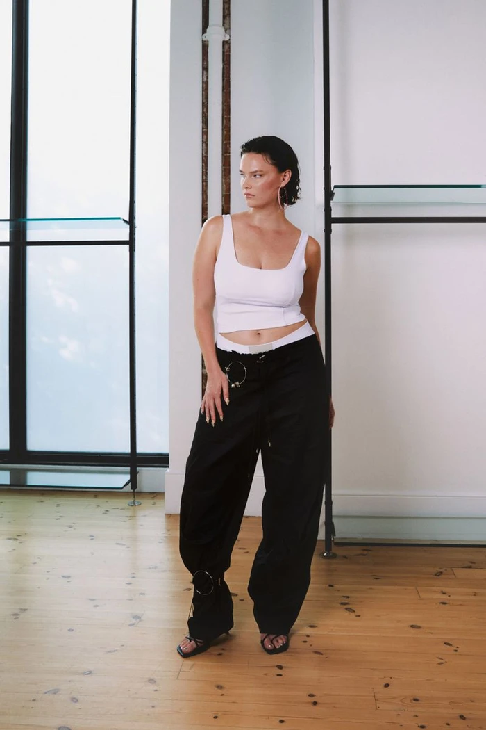 Model wearing a white tank top and black trousers standing in an interior space with wooden floors