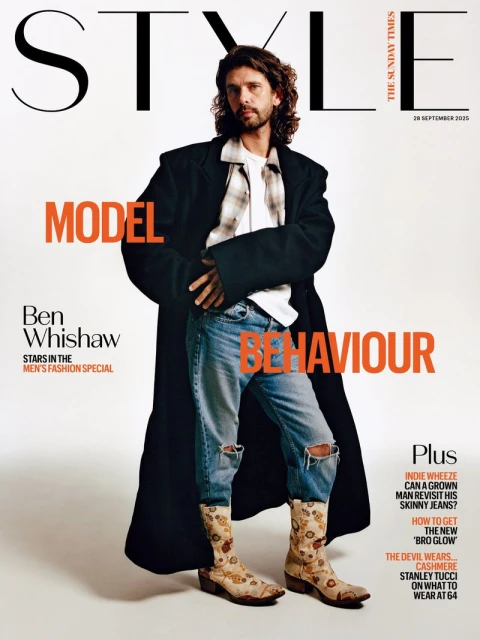 Fashion magazine cover featuring a male model wearing a long coat, jeans with knee rips, and floral patterned boots standing in a light studio.
