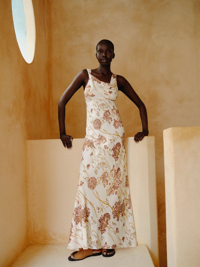 Elegant woman poses indoors, wearing a floral-patterned dress. Soft neutral light enhances the dress's detailed design and the indoor setting.