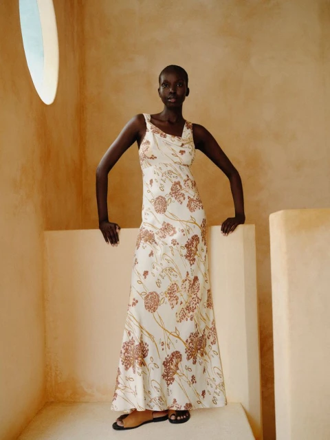 Elegant woman poses indoors, wearing a floral-patterned dress. Soft neutral light enhances the dress's detailed design and the indoor setting.