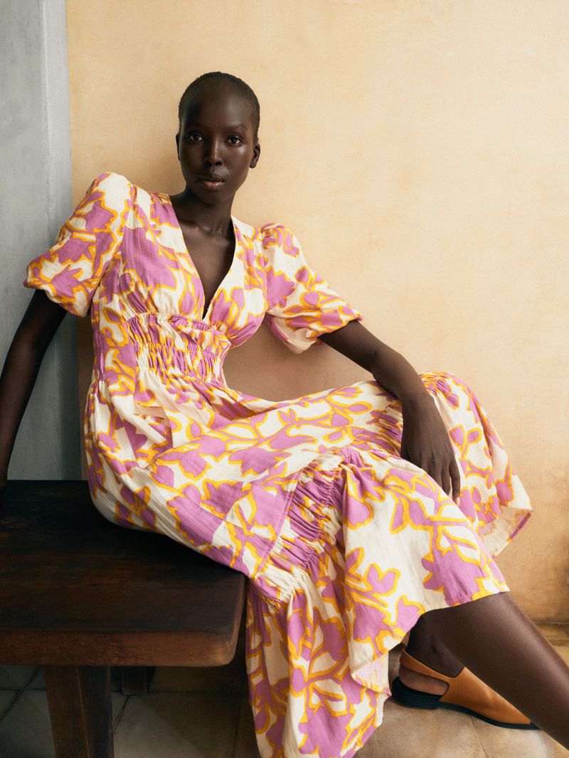 Fashion portrait of an adult woman in a pink and yellow leaf-print midi dress sitting on a wood bench in an indoor studio.