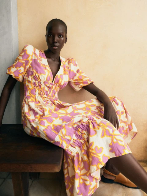 Fashion portrait of an adult woman in a pink and yellow leaf-print midi dress sitting on a wood bench in an indoor studio.