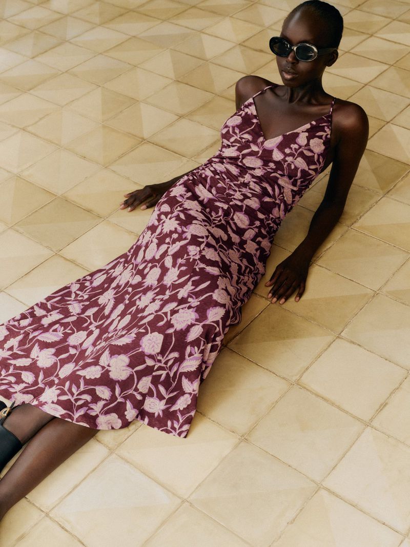 A woman in a floral dress and sunglasses lies on a tiled floor. The floral pattern on the dress is light pink and maroon.